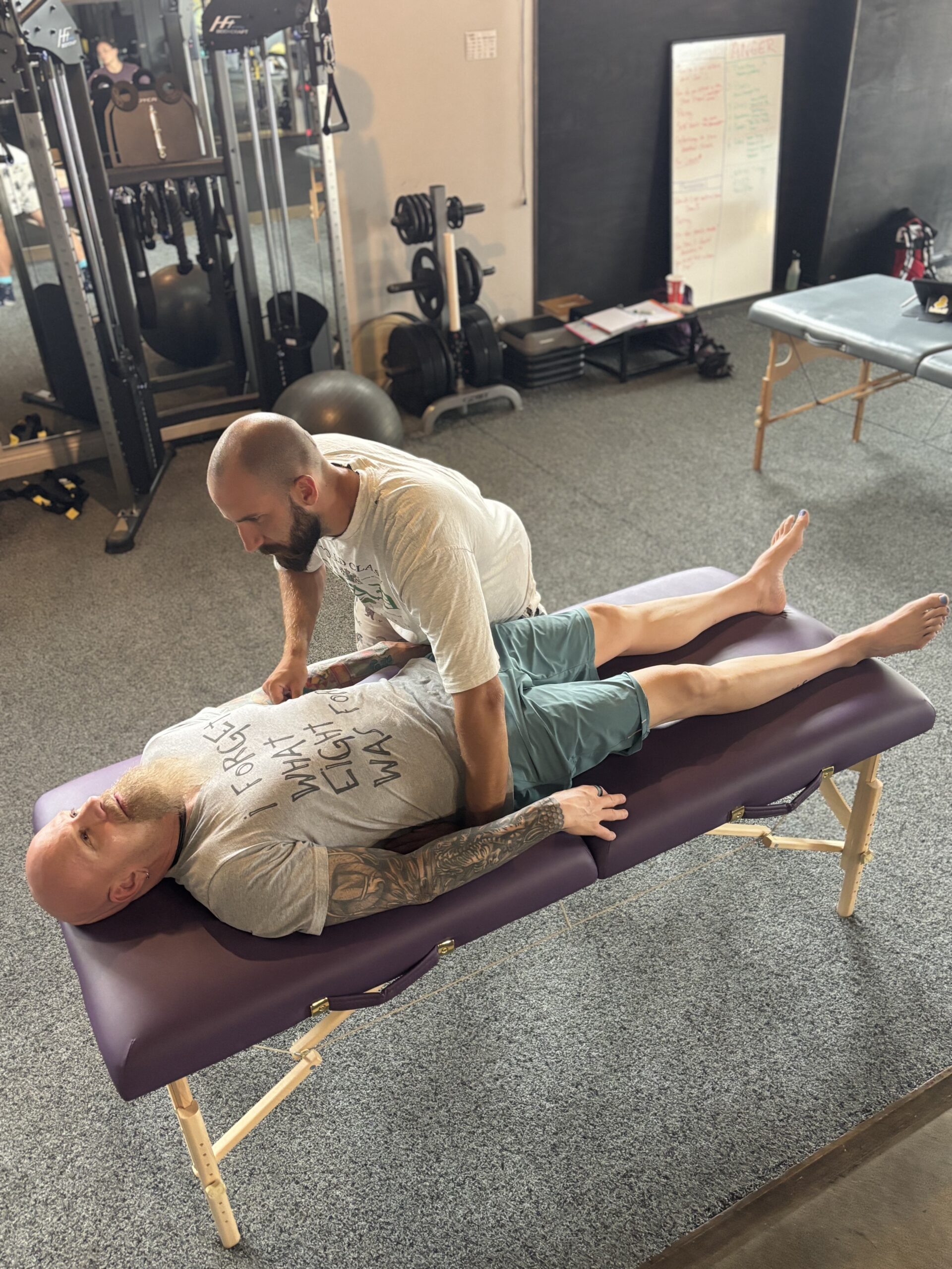 A person in a yellow sports shirt and green shorts performs squats on a stability ball while holding dumbbells, guided by a trainer.